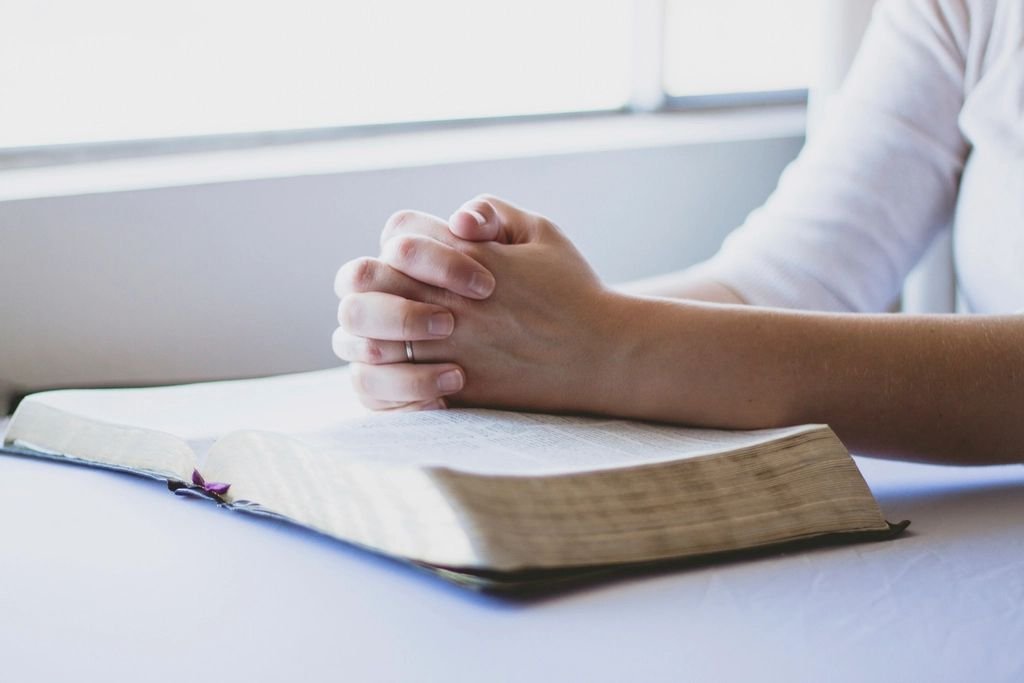 A woman's hands folded in prayer over an open Bible by a bright window – praying for my husband's success and protection
